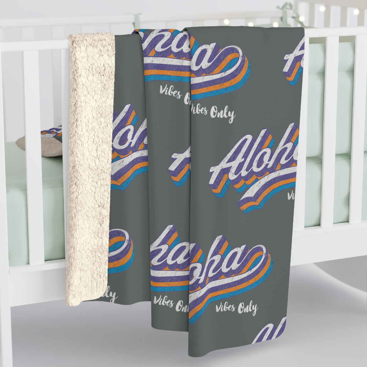 A baby crib with a dark gray blanket draped over the side, featuring a repeating 'Aloha vibes only' graphic in white, purple, orange, and blue, with a soft cream sherpa backing visible on the left.