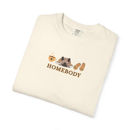 A folded off-white short-sleeve crewneck t-shirt with a graphic on the chest depicting a dog under a blanket, a coffee mug, a pair of house slippers, and the word 'HOMEBODY' in brown text.