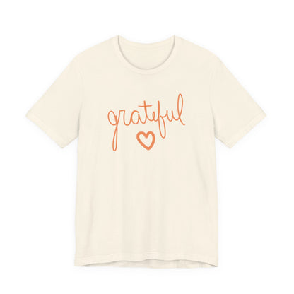 An off-white short-sleeve crewneck t-shirt featuring the word 'grateful' in orange cursive script and a matching orange heart outline on the chest, displayed against a solid white background.
