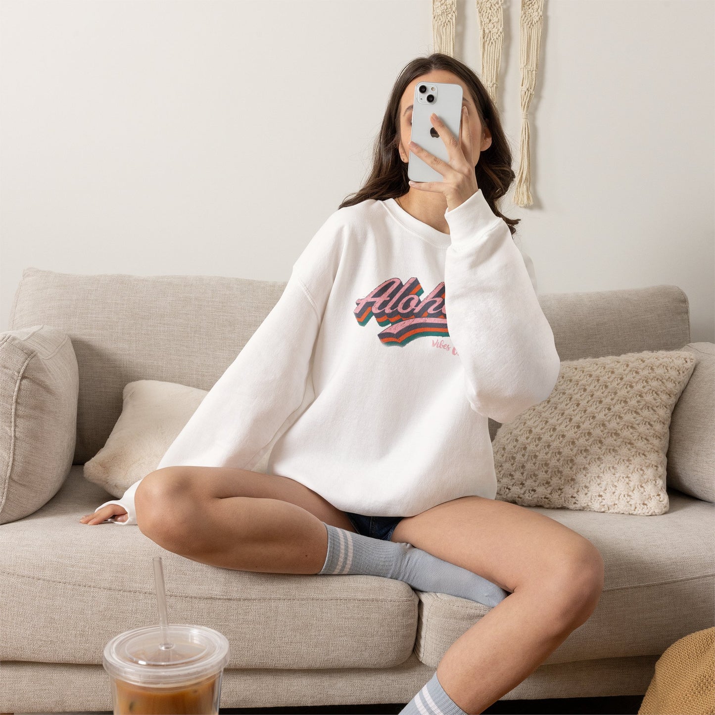 A woman sitting on a beige couch takes a mirror selfie while wearing a white crewneck sweatshirt with a colorful retro 'Aloha' graphic on the chest.