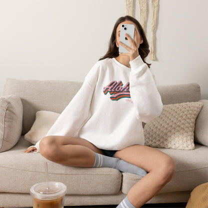 A woman sitting on a beige couch takes a mirror selfie while wearing a white crewneck sweatshirt with a colorful retro 'Aloha' graphic on the chest.