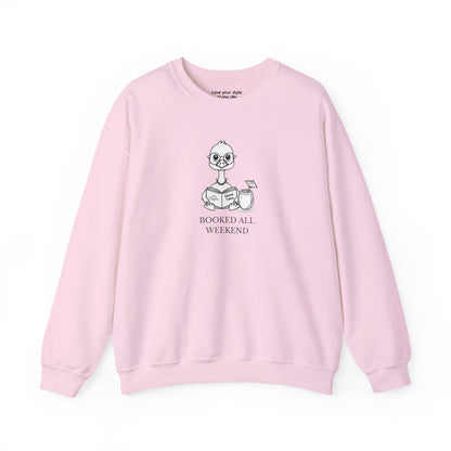 Pink sweatshirt with graphic print of a cartoon nene goose sitting reading a book with a cocktail next to it with text 'BOOKED ALL WEEKEND'