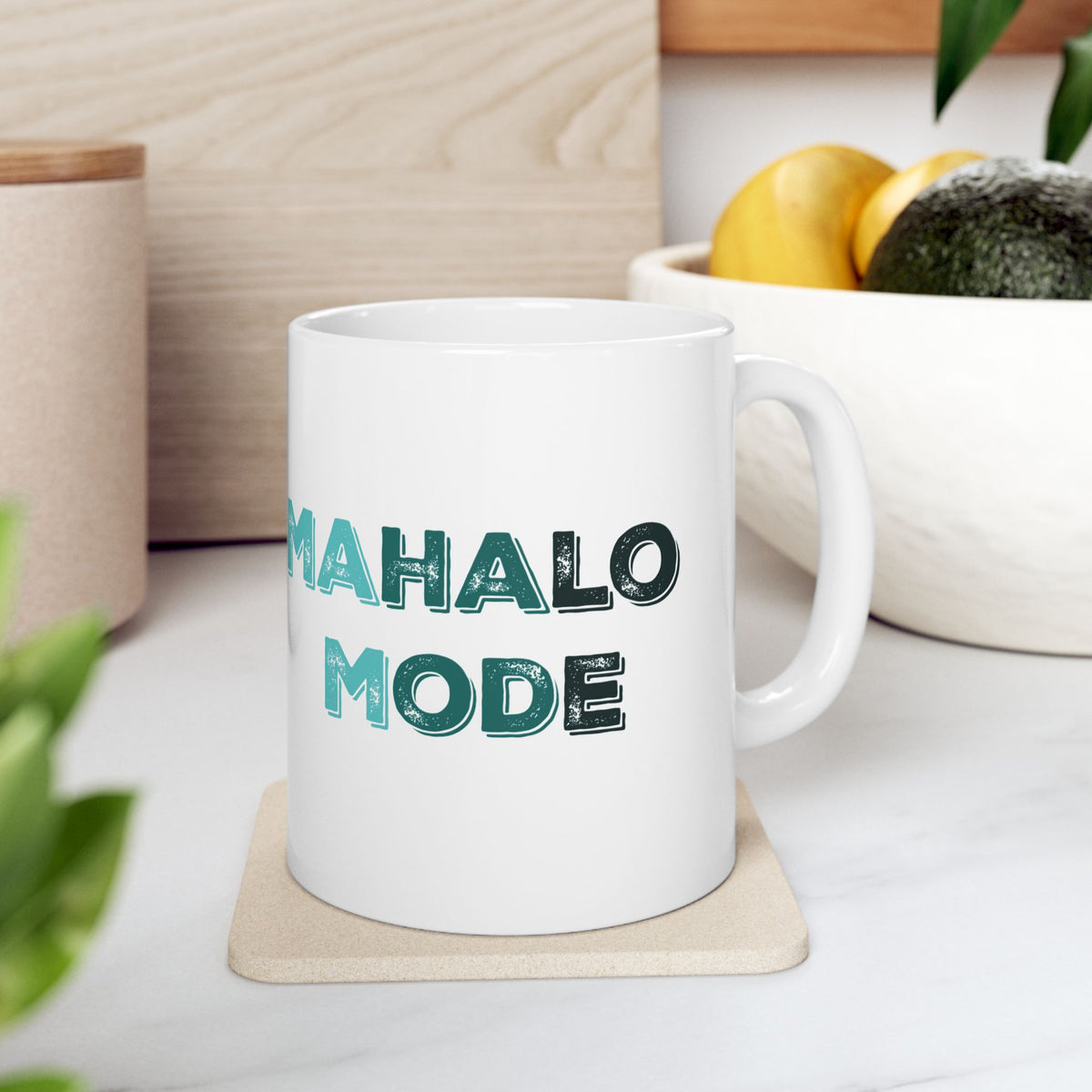 A white ceramic mug with the words 'MAHALO MODE' printed in a textured teal font, resting on a beige coaster on a kitchen counter with a bowl of fruit in the background.