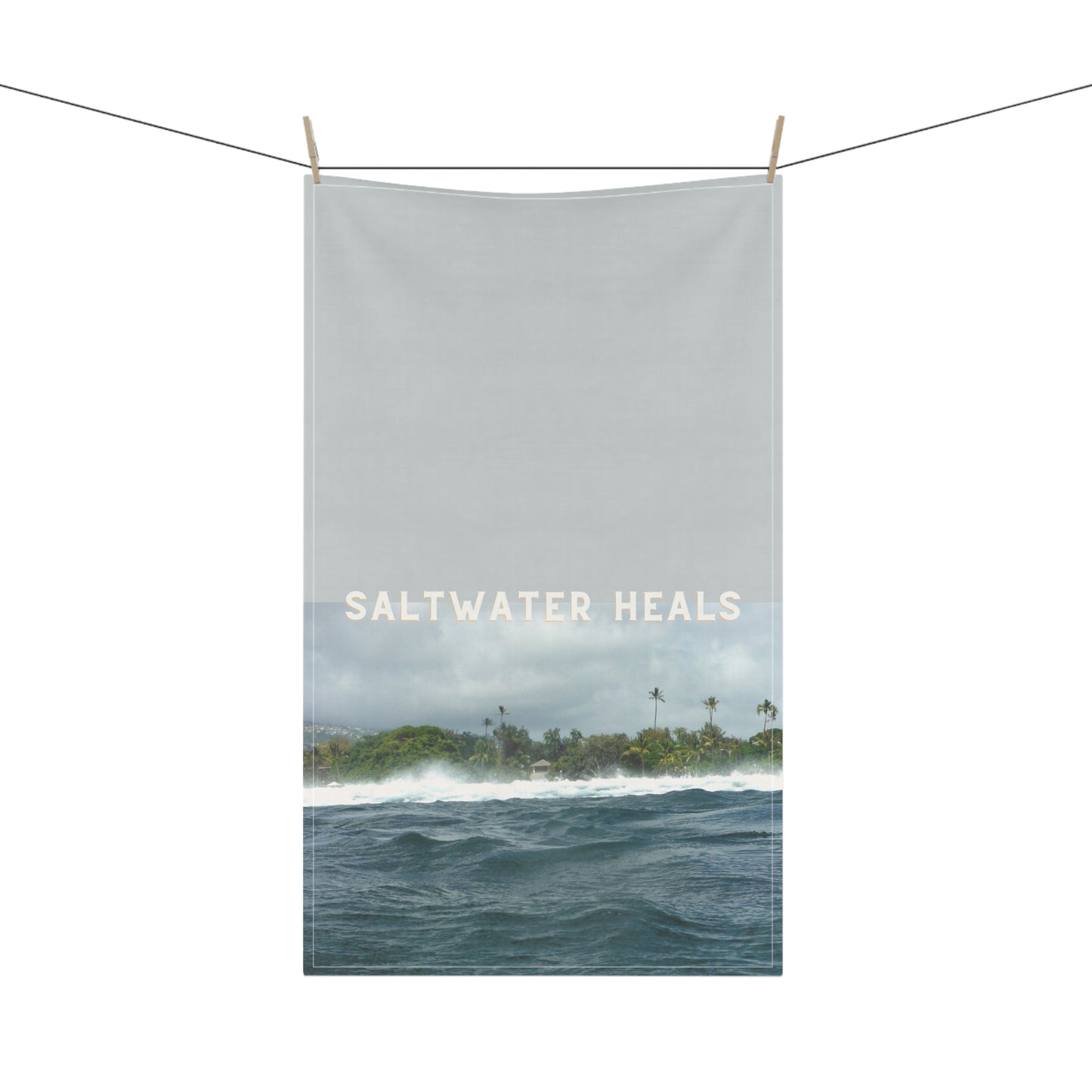 A rectangular tea towel hanging from a string with wooden clips against a white background. The artwork shows a seascape with dark blue ocean waves, a distant tropical shoreline with palm trees, and a pale sky.