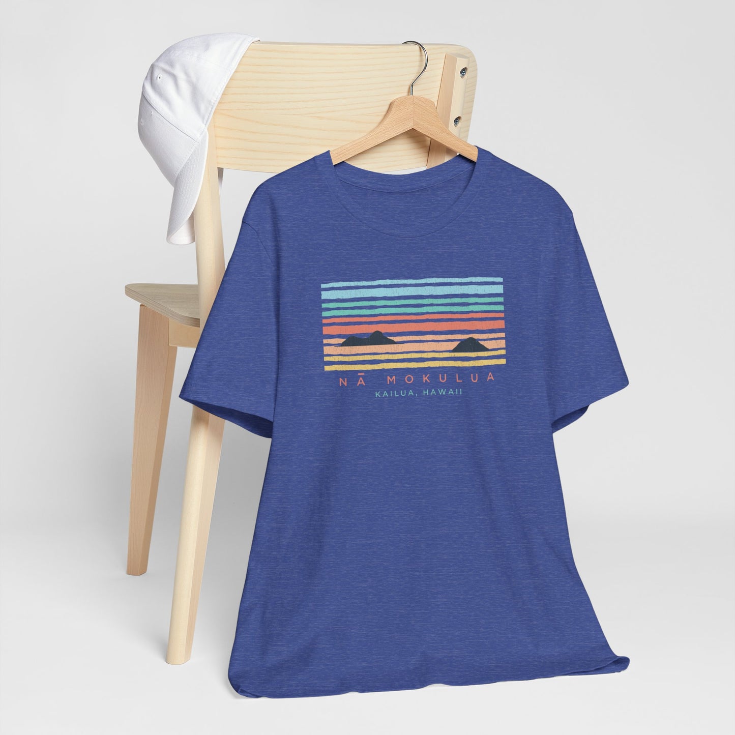 A hanging blue heathered t-shirt with a colorful retro sunset graphic and the text 'NÁ MOKULUA KAILUA, HAWAII' on the front, hanging from a wood chair.