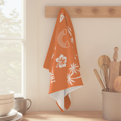 An orange kitchen towel with white tropical and Hawaiian-themed prints hangs from a wooden peg rack on a beige wall in a bright kitchen setting.