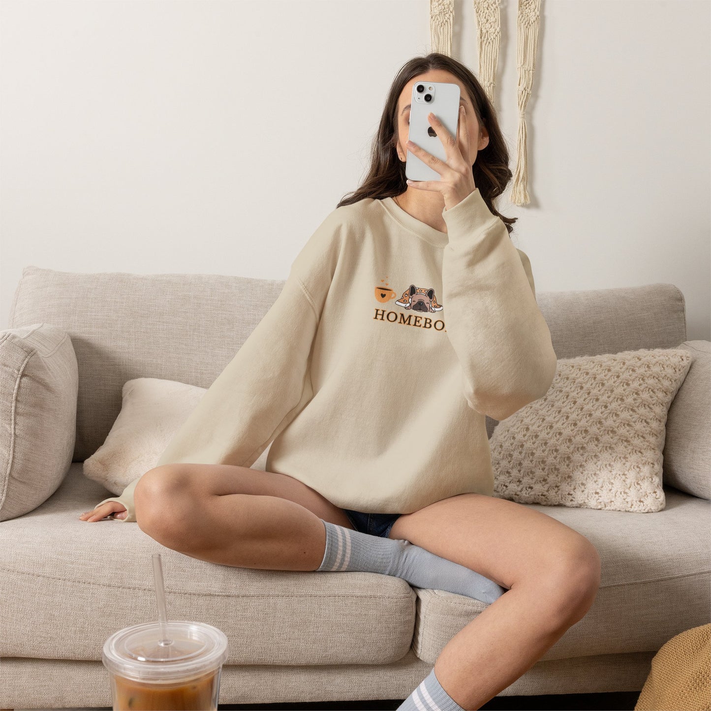 Person wearing a beige crewneck sweatshirt featuring a central graphic of a dog under a blanket, house slippers, coffee cups and the word 'HOMEBODY' printed below it sitting on a couch taking a picture.