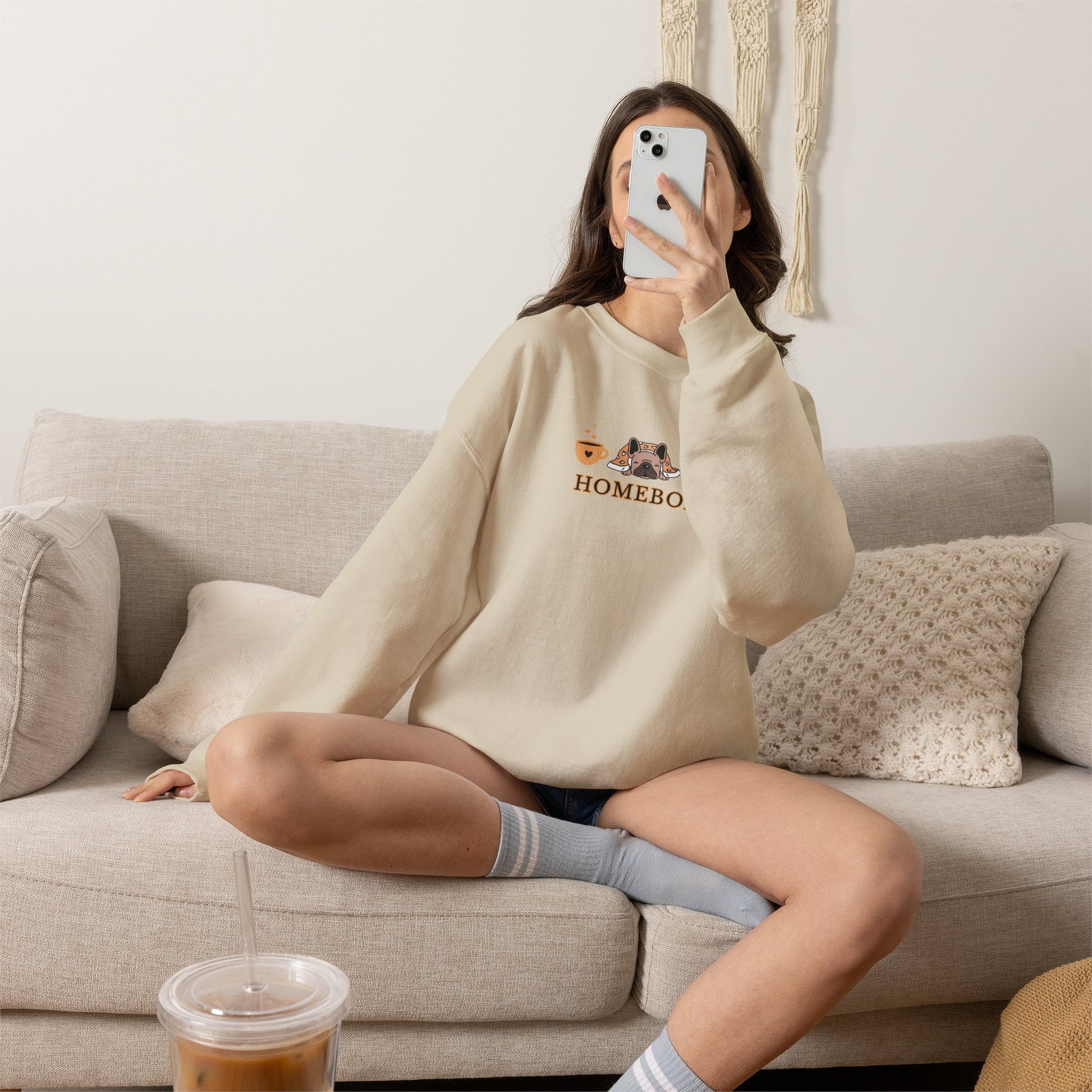 Person wearing a beige crewneck sweatshirt featuring a central graphic of a dog under a blanket, house slippers, coffee cups and the word 'HOMEBODY' printed below it sitting on a couch taking a picture.