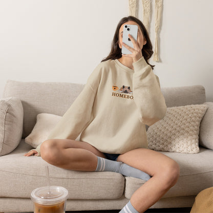 Person wearing a beige crewneck sweatshirt featuring a central graphic of a dog under a blanket, house slippers, coffee cups and the word 'HOMEBODY' printed below it sitting on a couch taking a picture.
