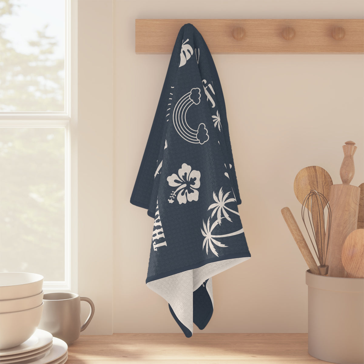 A navy blue kitchen towel with white tropical and beach-themed prints hangs from a wooden peg on a kitchen wall. The towel features white palm trees, a rainbow, a hibiscus flower, and the word 'Thankful.