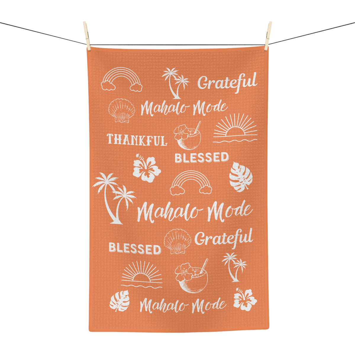 An orange rectangular kitchen towel with white tropical-themed text and illustrations, hanging from a clothesline with wooden clips against a white background.