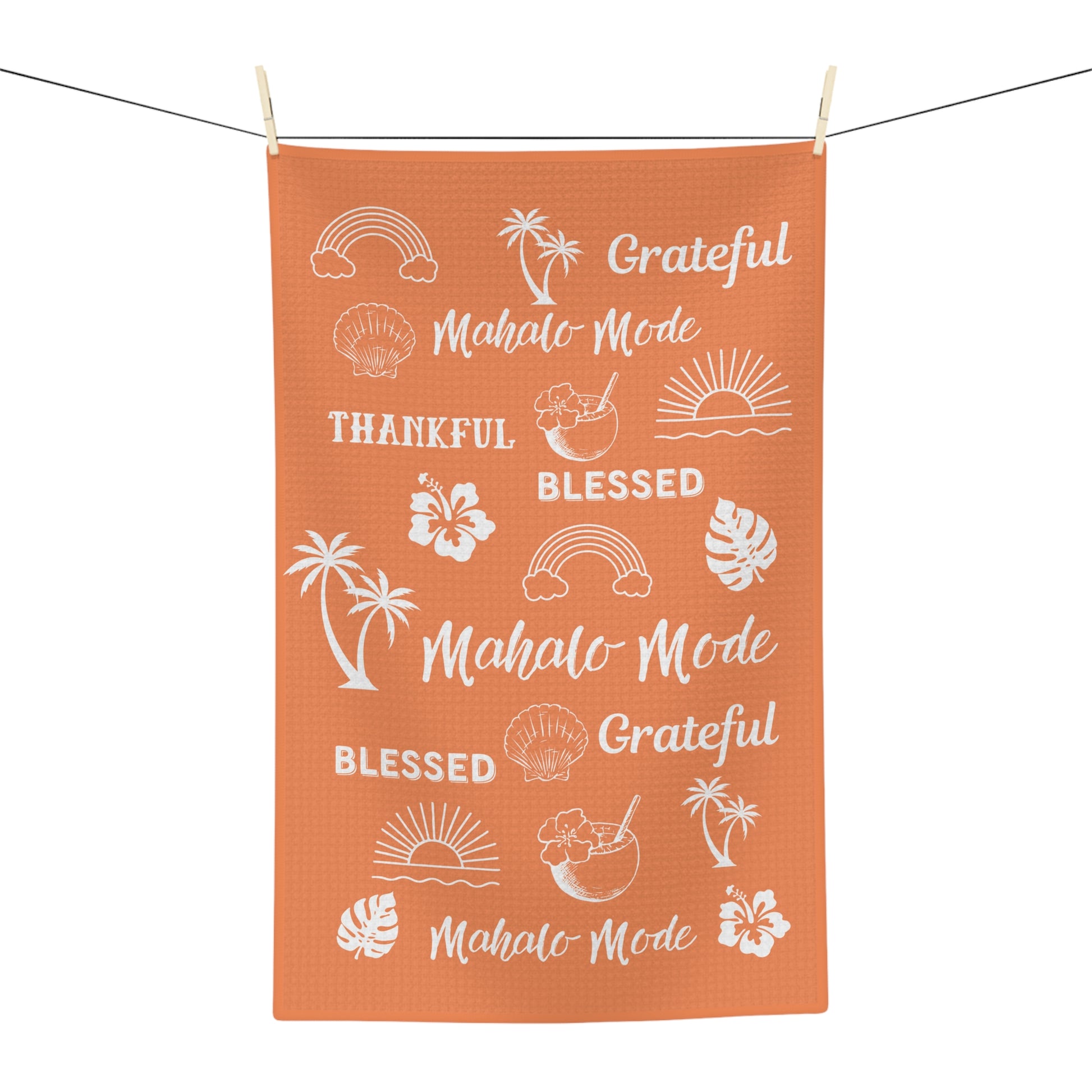 An orange rectangular kitchen towel with white tropical-themed text and illustrations, hanging from a clothesline with wooden clips against a white background.