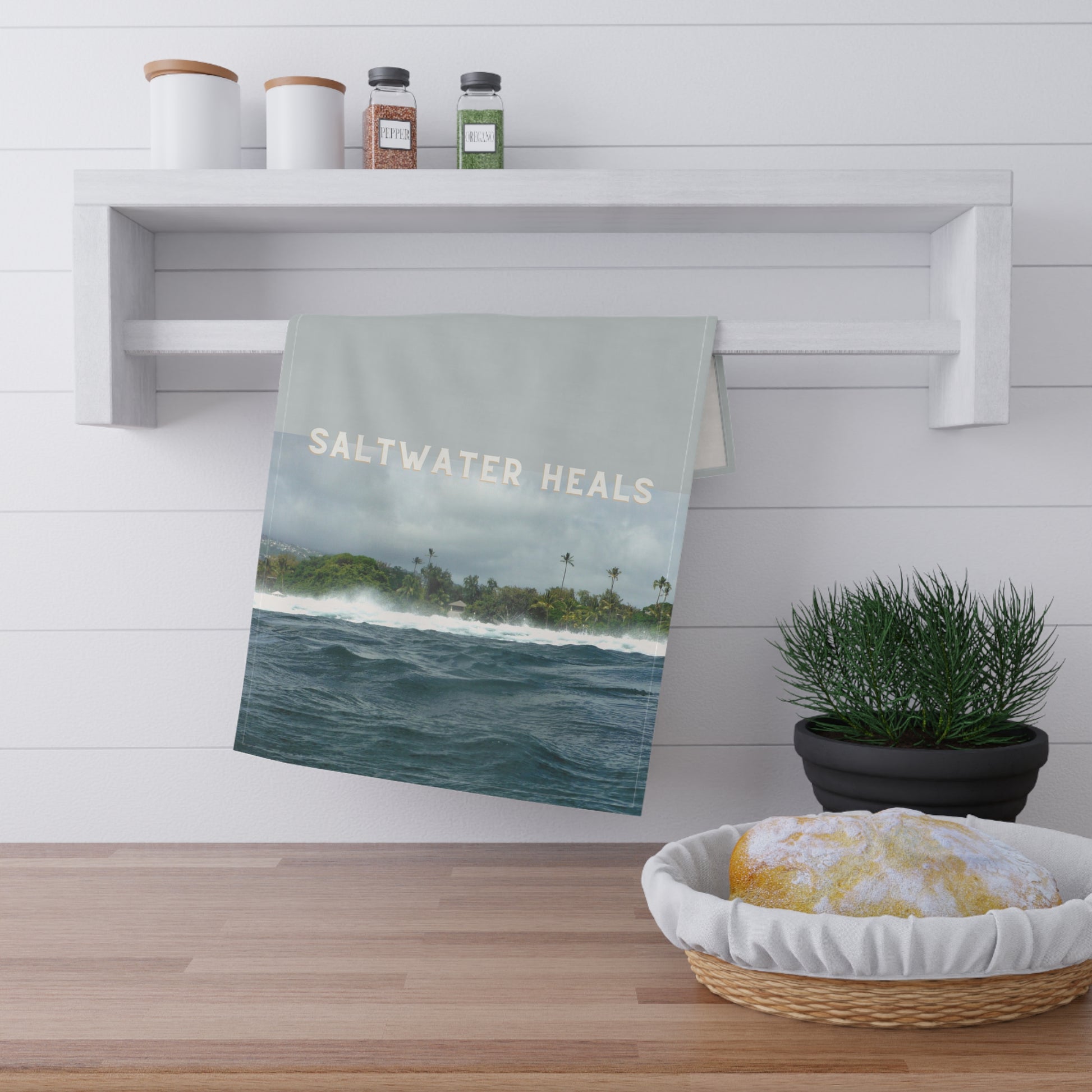 A kitchen towel with a scenic ocean print and the words 'SALTWATER HEALS' hanging from a white wall-mounted rack in a bright kitchen setting.
