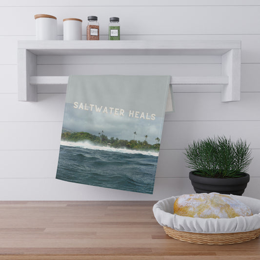A kitchen towel with a scenic ocean print and the words 'SALTWATER HEALS' hanging from a white wall-mounted rack in a bright kitchen setting.