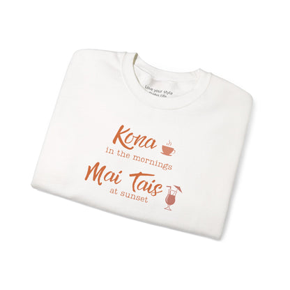 A folded white crewneck sweatshirt with orange text reading 'Kona in the mornings Mai Tais at sunset' and small coffee cup and cocktail glass illustrations, shown on a white background.
