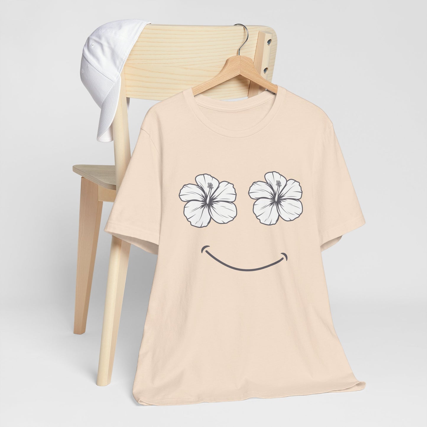 A light beige short-sleeve t-shirt on a wooden hanger, featuring a graphic of two white hibiscus flowers for eyes and a black smiley mouth. The shirt is displayed on a wooden chair with a white t-shirt draped over the back against a light gray background.