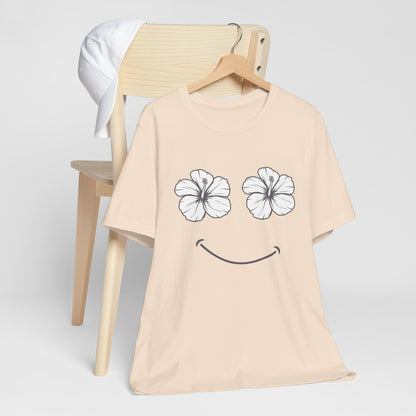 A light beige short-sleeve t-shirt on a wooden hanger, featuring a graphic of two white hibiscus flowers for eyes and a black smiley mouth. The shirt is displayed on a wooden chair with a white t-shirt draped over the back against a light gray background.