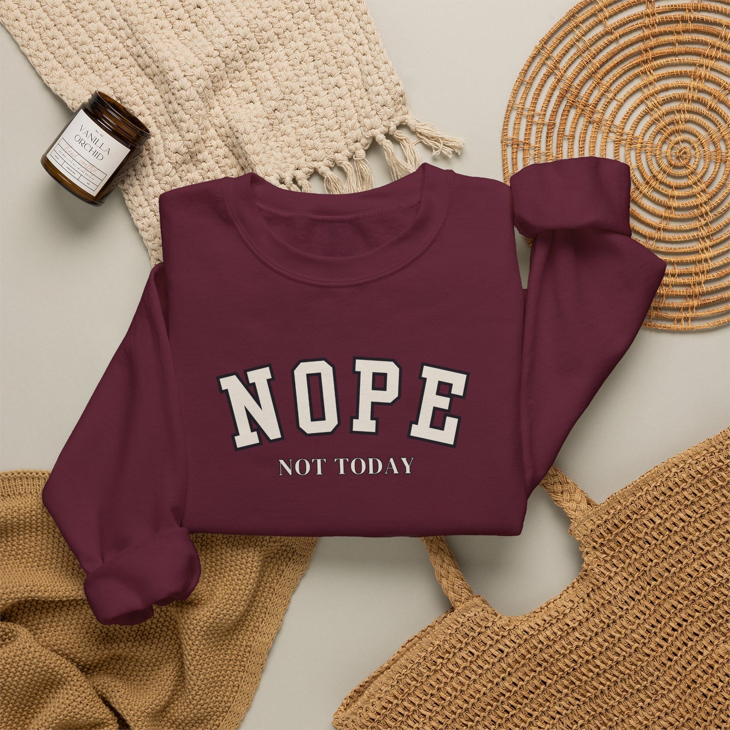 A folded long-sleeve sweatshirt in deep burgundy with the words 'NOPE NOT TODAY' printed in white varsity-style lettering, styled in a flat lay with a woven blanket, a small candle jar, and a wicker tray on a light neutral background.