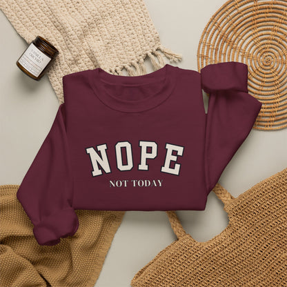A folded long-sleeve sweatshirt in deep burgundy with the words 'NOPE NOT TODAY' printed in white varsity-style lettering, styled in a flat lay with a woven blanket, a small candle jar, and a wicker tray on a light neutral background.
