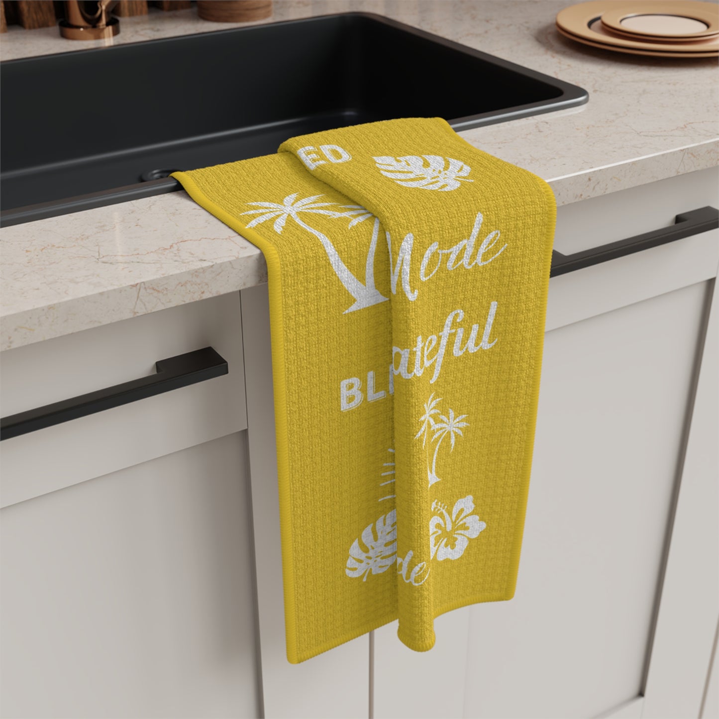 A bright yellow kitchen towel with white tropical palm tree and hibiscus flower prints hangs over the edge of a black sink in a modern kitchen setting.
