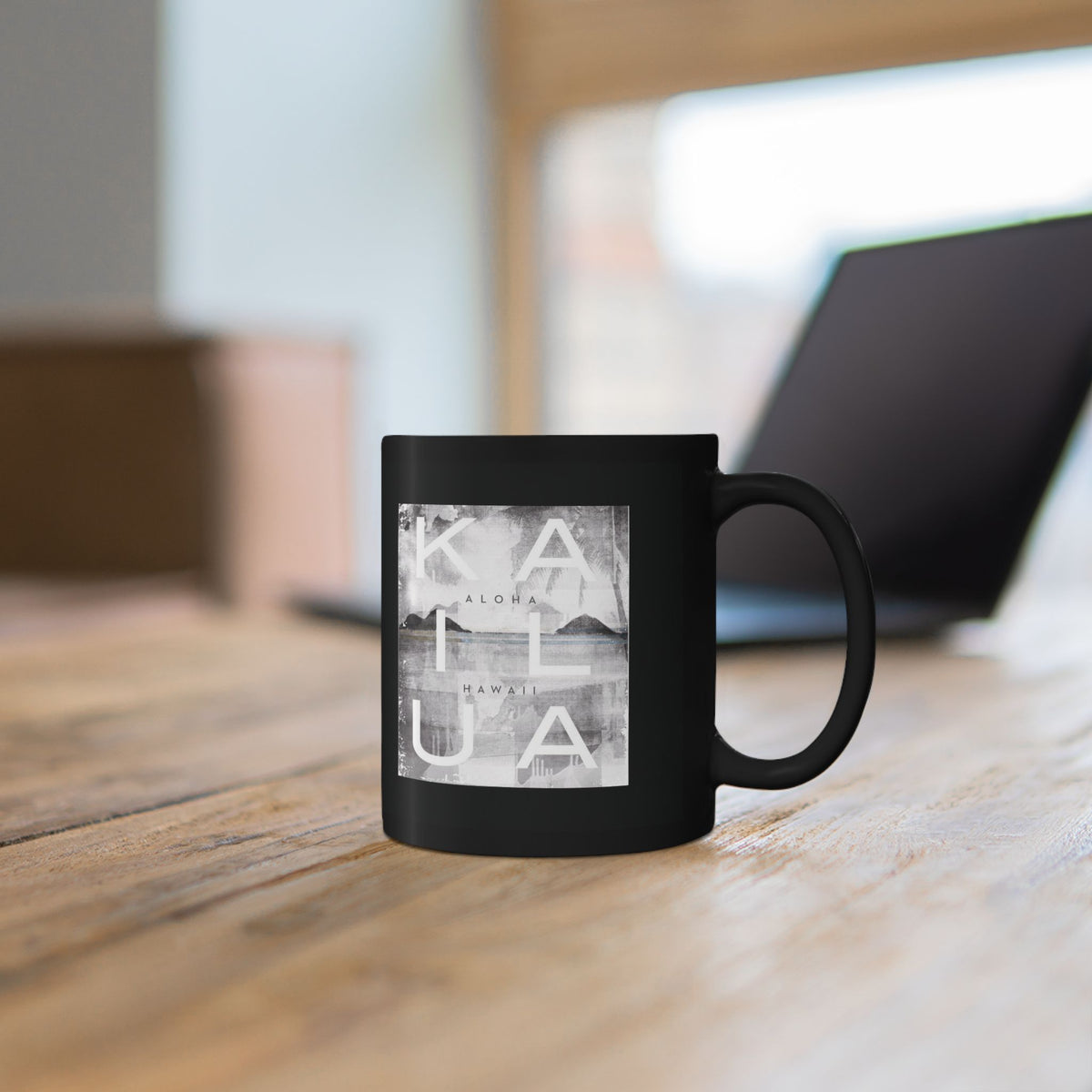 A black ceramic mug with a square white and gray graphic print reading 'KAILUA' and smaller text, sitting on a wooden table with a blurred laptop in the background.