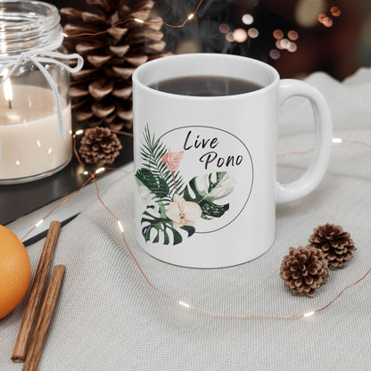 A white ceramic mug with a tropical leaf graphic and the words 'Live Pono' printed on it, filled with black coffee and styled on a tablecloth with pinecones, a candle, cinnamon sticks, and fairy lights.
