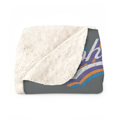 A folded throw blanket featuring a plush cream sherpa fleece backing and a dark gray printed front with a colorful retro-style aloha vibes only graphic.