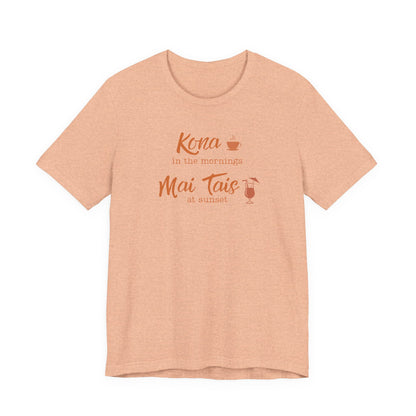 A heather peach short-sleeve t-shirt with a coral orange graphic on the chest that reads 'Kona in the mornings Mai Tais at sunset' with small coffee and cocktail illustrations, displayed on a clean white background.