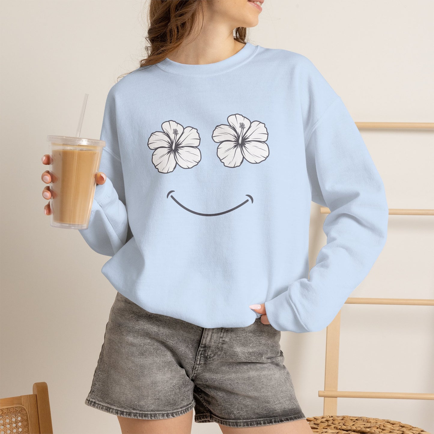 A woman wearing a light blue Happy Hibiscus Sweatshirt featuring a cheerful hibiscus flower smile design inspired by island life and aloha style holding a coffee