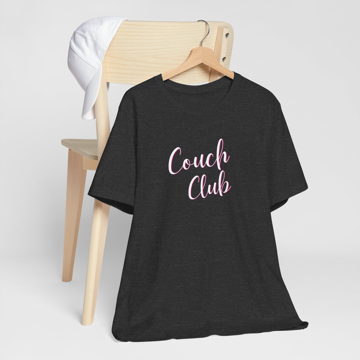 A black short-sleeve t-shirt on a wooden hanger, styled in front of a light wood chair with a white cap. The t-shirt features the text 'Couch Club' in white script.