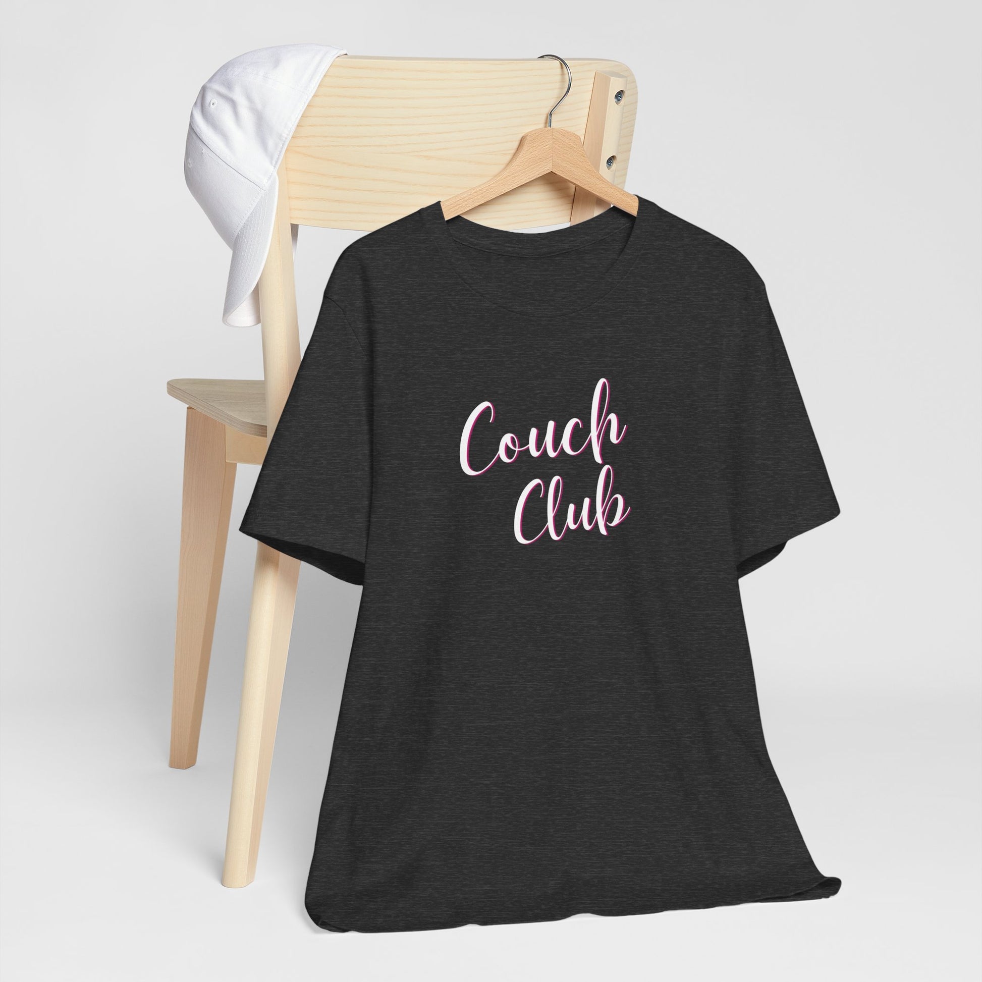 A black short-sleeve t-shirt on a wooden hanger, styled in front of a light wood chair with a white cap. The t-shirt features the text 'Couch Club' in white script.