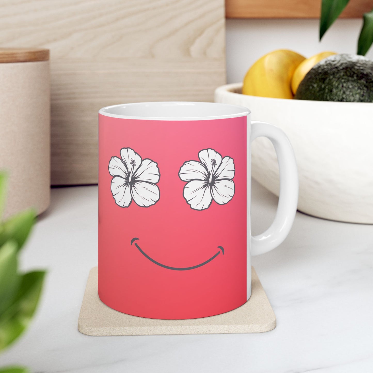 A pink ceramic mug with a white handle and interior, featuring a minimalist smiley face made of two white hibiscus flowers for eyes and a curved smile. The mug sits on a square coaster on a white marble-like surface, with a wooden block, bowl of fruit, and green leaves in the background.