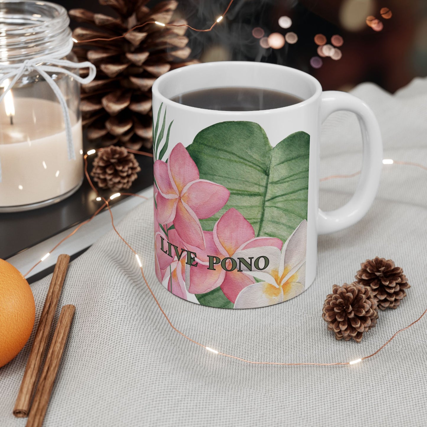 A white ceramic mug with a tropical floral print featuring pink plumeria flowers, green leaves, and the text 'LIVE PONO', filled with black coffee and styled on a table with a candle, pinecones, cinnamon sticks, and fairy lights.