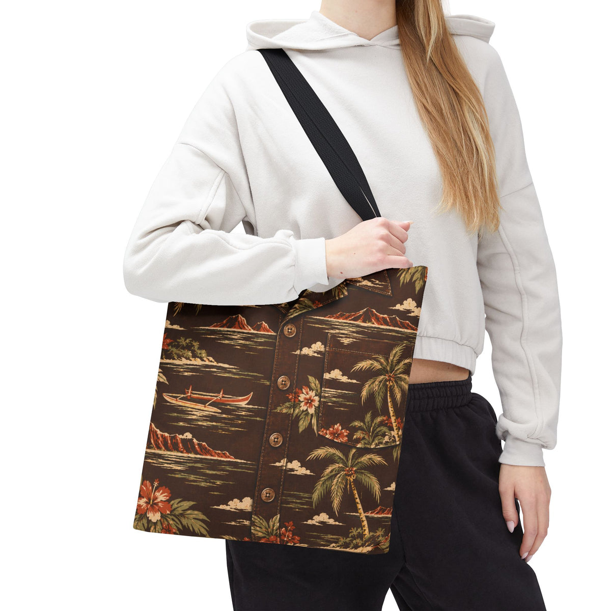 A woman wearing a white hoodie and black sweatpants carries a brown button-up shirt-style tote bag with a tropical print featuring palm trees, canoes, mountains, and flowers. The bag has a black shoulder strap and is shown against a plain white background.