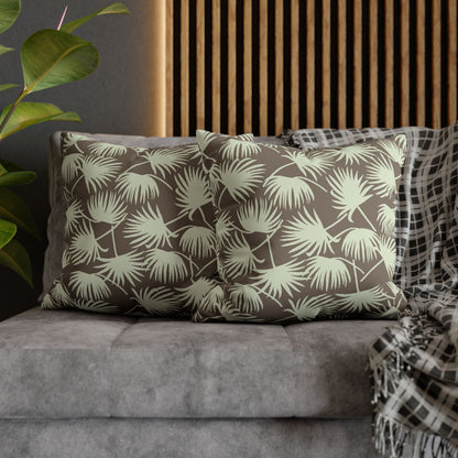 Two square throw pillows with a palm leaf pattern in olive green and taupe, styled on a gray sofa with a plaid throw blanket and a wooden slat wall background.