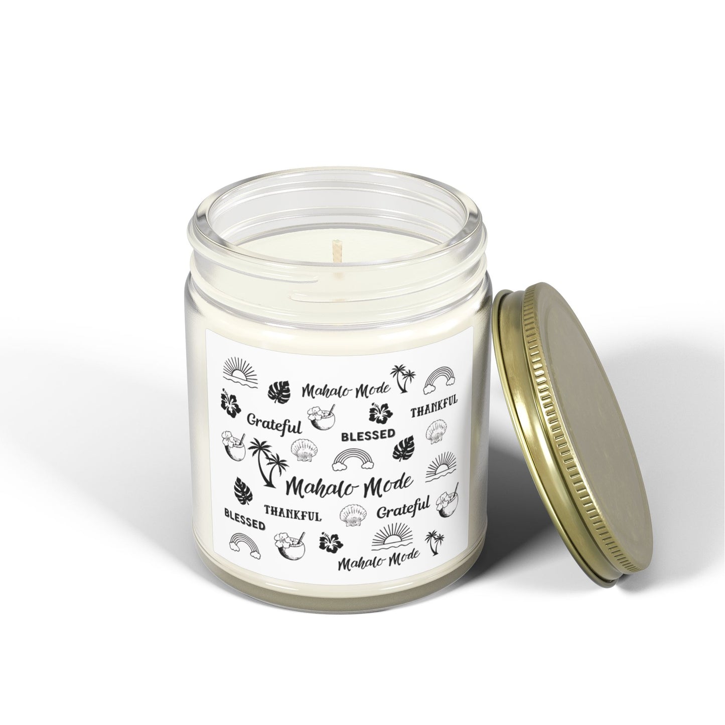 A glass jar candle with a gold metal lid and a white paper label featuring black tropical-themed illustrations and text like 'Mahalo Mode,' 'Grateful,' and 'Blessed,' shown on a clean white background.
