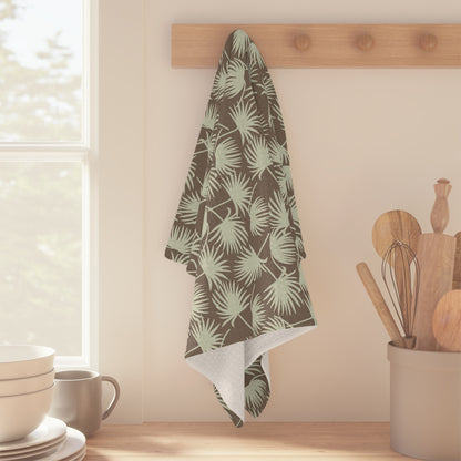 A kitchen towel with a green palm leaf pattern hanging from a wooden peg on a beige wall, with kitchen utensils and a window in the background.