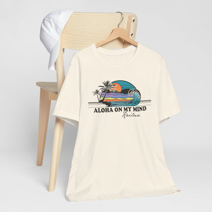 An off-white short-sleeve graphic t-shirt hanging on a wooden hanger from a light wood chair, with a white cap draped over the backrest. The shirt features a colorful retro sunset beach graphic with palm trees and the text 'ALOHA ON MY MIND' and 'Kailua'.