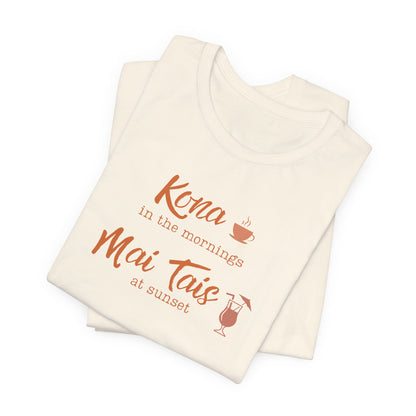 A folded cream-colored t-shirt with orange text reading 'Kona in the mornings Mai Tais at sunset' and small orange graphic illustrations of a coffee cup and a cocktail glass, shown on a plain white background.