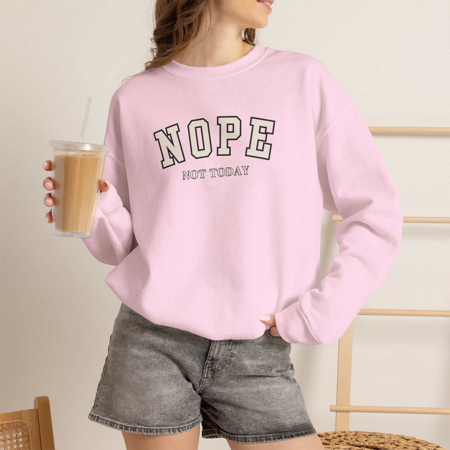 A woman wearing a light pink crewneck sweatshirt with the text 'NOPE NOT TODAY' printed on the front in black and white, paired with gray denim shorts and holding an iced coffee.