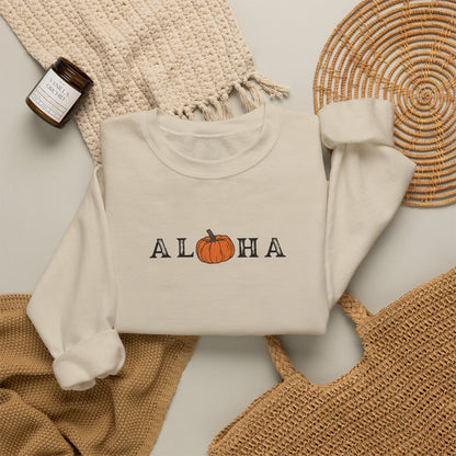 A sand crewneck sweatshirt with 'ALOHA' and a pumpkin graphic printed on the chest, styled in a flat lay with a woven blanket, a small candle, and a wicker tray.