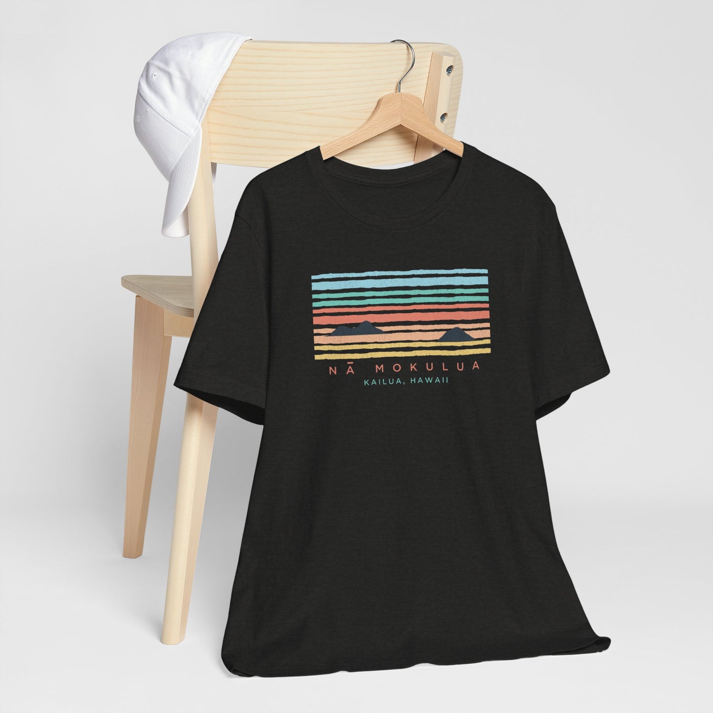 A hanging black t-shirt with a colorful retro sunset graphic and the text 'NÁ MOKULUA KAILUA, HAWAII' on the front, hanging from a wood chair.