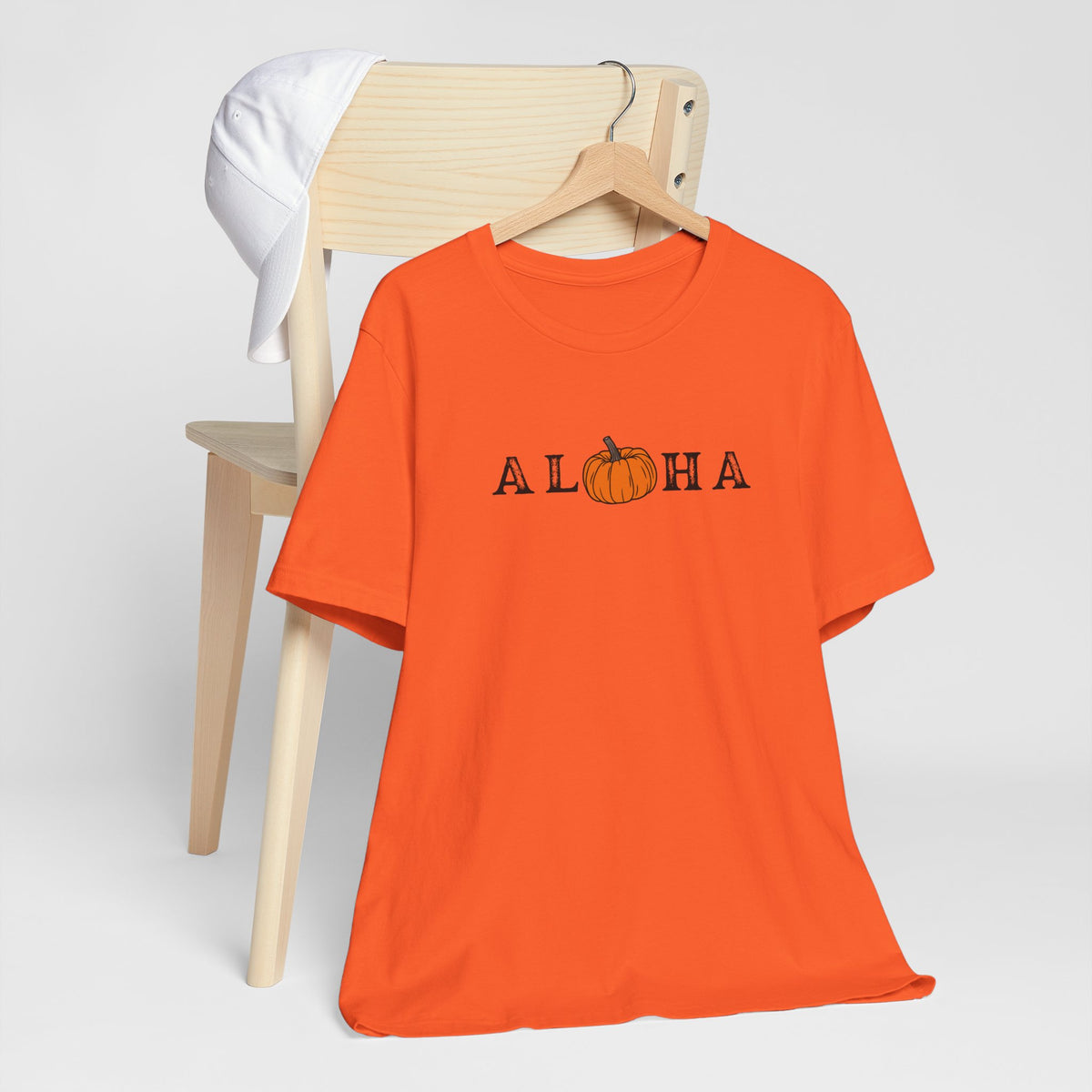An orange short-sleeve t-shirt on a wooden hanger, featuring the word 'ALOHA' with a pumpkin graphic replacing the 'O'. A light wood chair and a white towel are in the background against a light gray studio backdrop.
