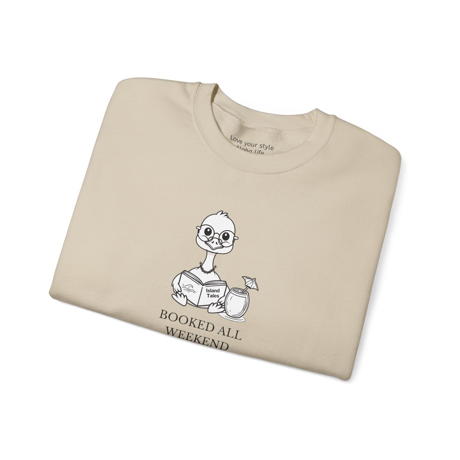 A folded sand crewneck sweatshirt featuring a cartoon nene goose character reading a book with the text 'BOOKED ALL WEEKEND' underneath, displayed on a clean white background.
