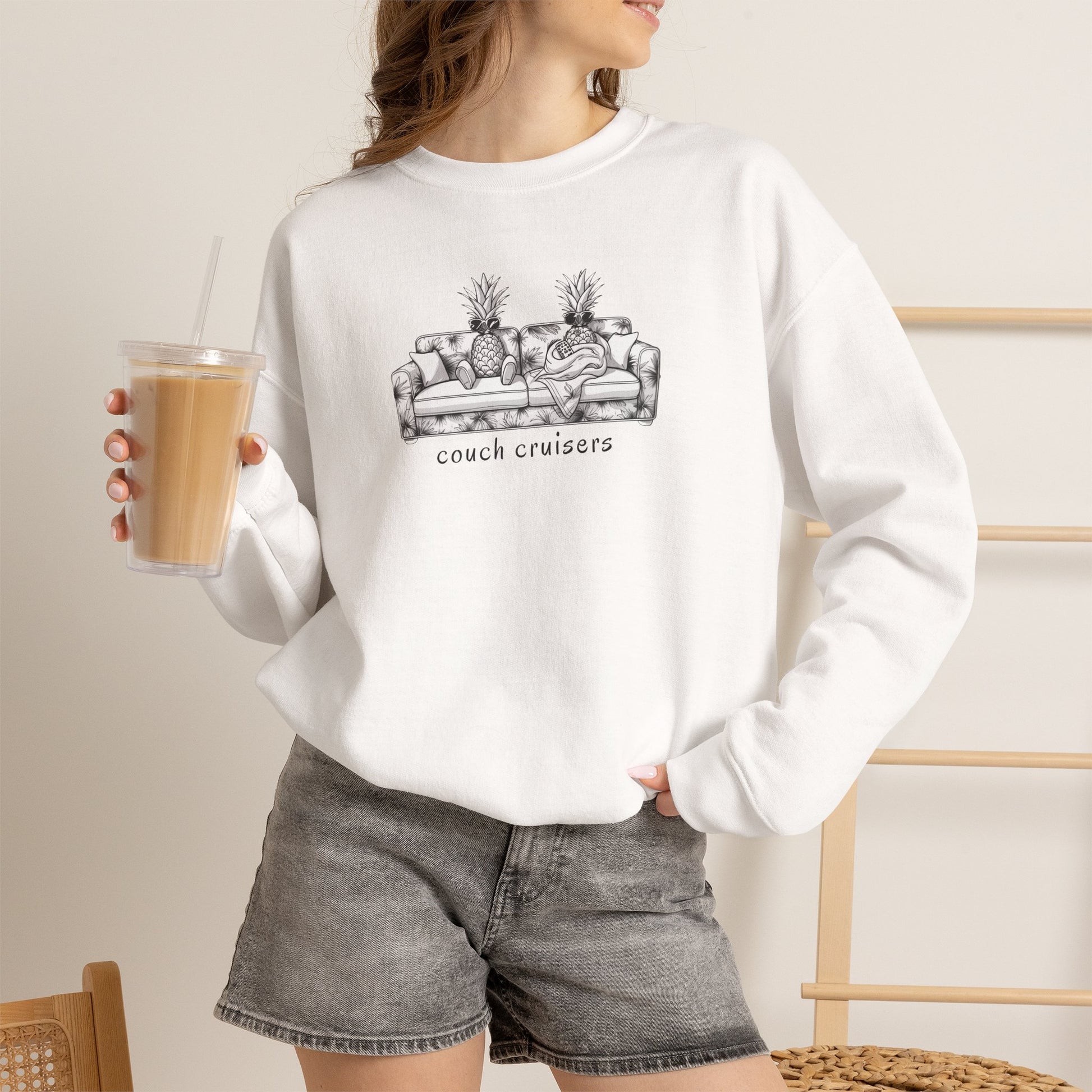 A woman wearing a white crewneck sweatshirt with a black line art graphic of a couch with pineapples and the text 'couch cruisers', paired with gray denim shorts, holding an iced coffee in a cafe setting.
