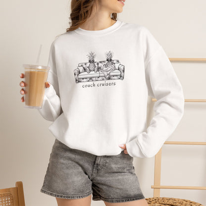 A woman wearing a white crewneck sweatshirt with a black line art graphic of a couch with pineapples and the text 'couch cruisers', paired with gray denim shorts, holding an iced coffee in a cafe setting.