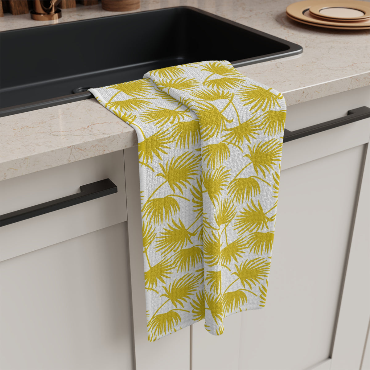 A kitchen towel with a yellow palm leaf pattern on a white background, draped over the edge of a black sink in a modern kitchen setting.