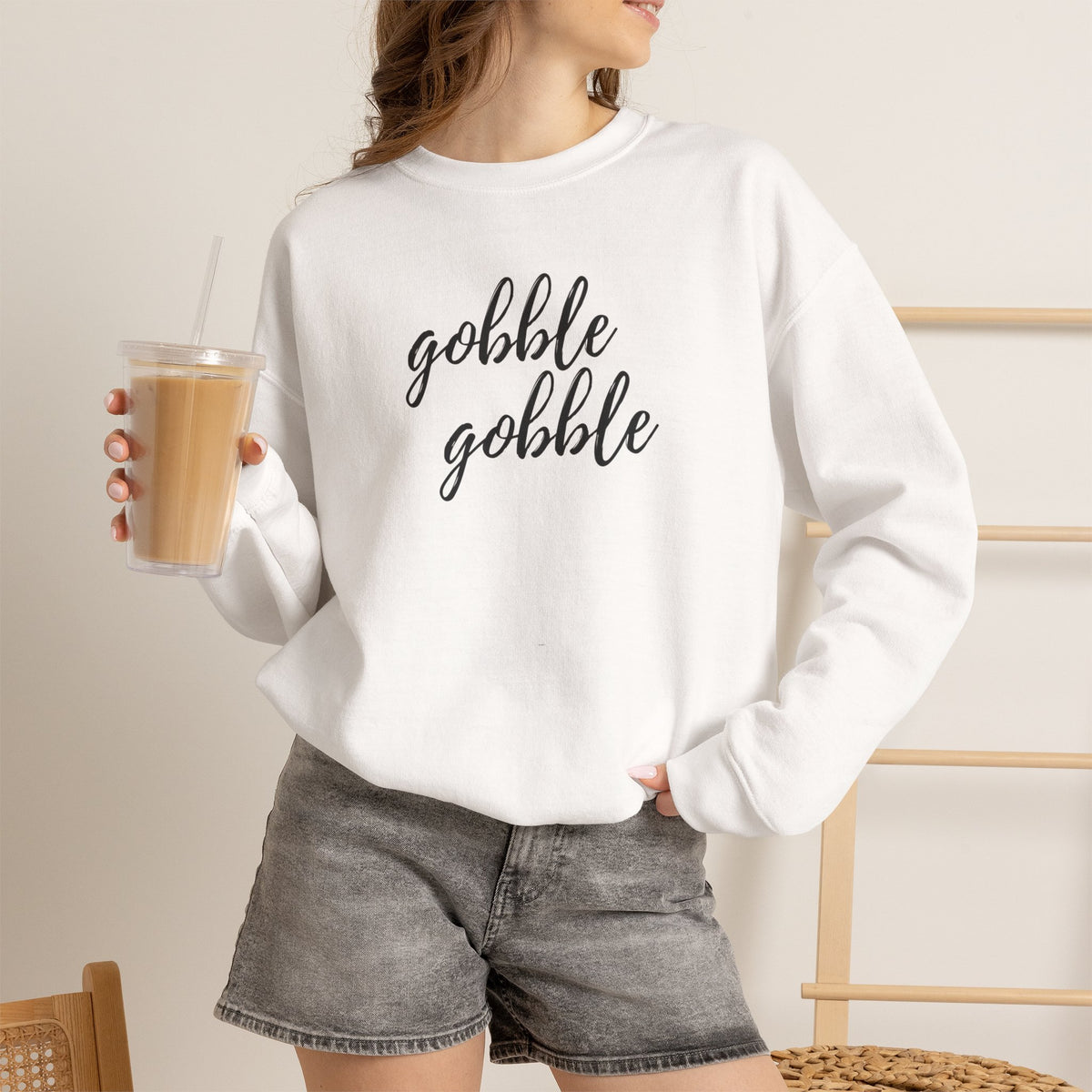A woman wearing a white crewneck sweatshirt with the phrase 'gobble gobble' printed in black script across the chest, paired with gray denim shorts and holding an iced coffee.