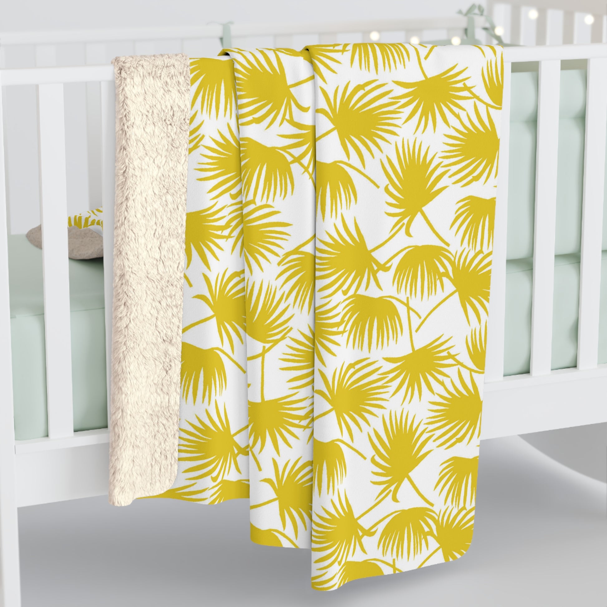 A throw blanket draped over a white crib, featuring a bold yellow palm leaf pattern on a white background with a soft cream sherpa fleece backing.