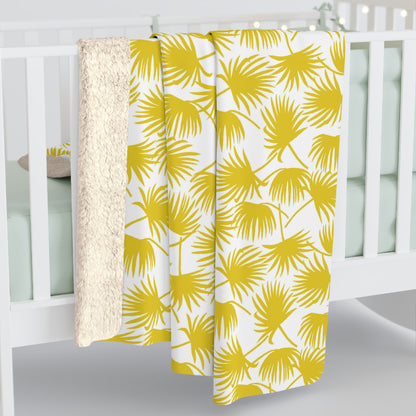 A throw blanket draped over a white crib, featuring a bold yellow palm leaf pattern on a white background with a soft cream sherpa fleece backing.